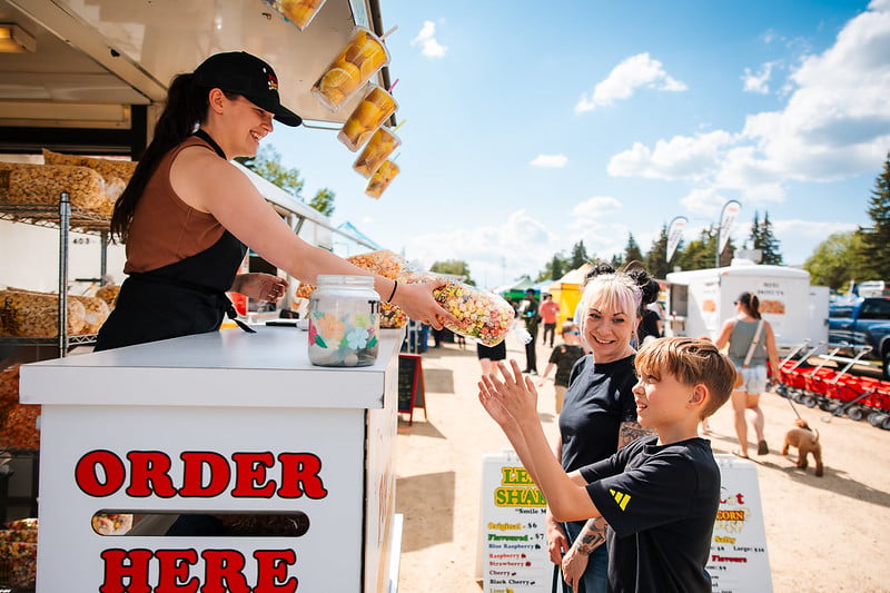 Vendor at Lacombe Market handing a colourful bag of popcorn to a young boy.